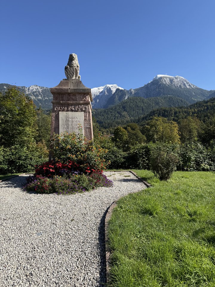 A stone war memorial topped with a carved lion, inscribed with dates 1939-1945, surrounded by red and purple flowers and a gravel path, with green forested mountains and snow-capped peaks in the background under a clear sky.