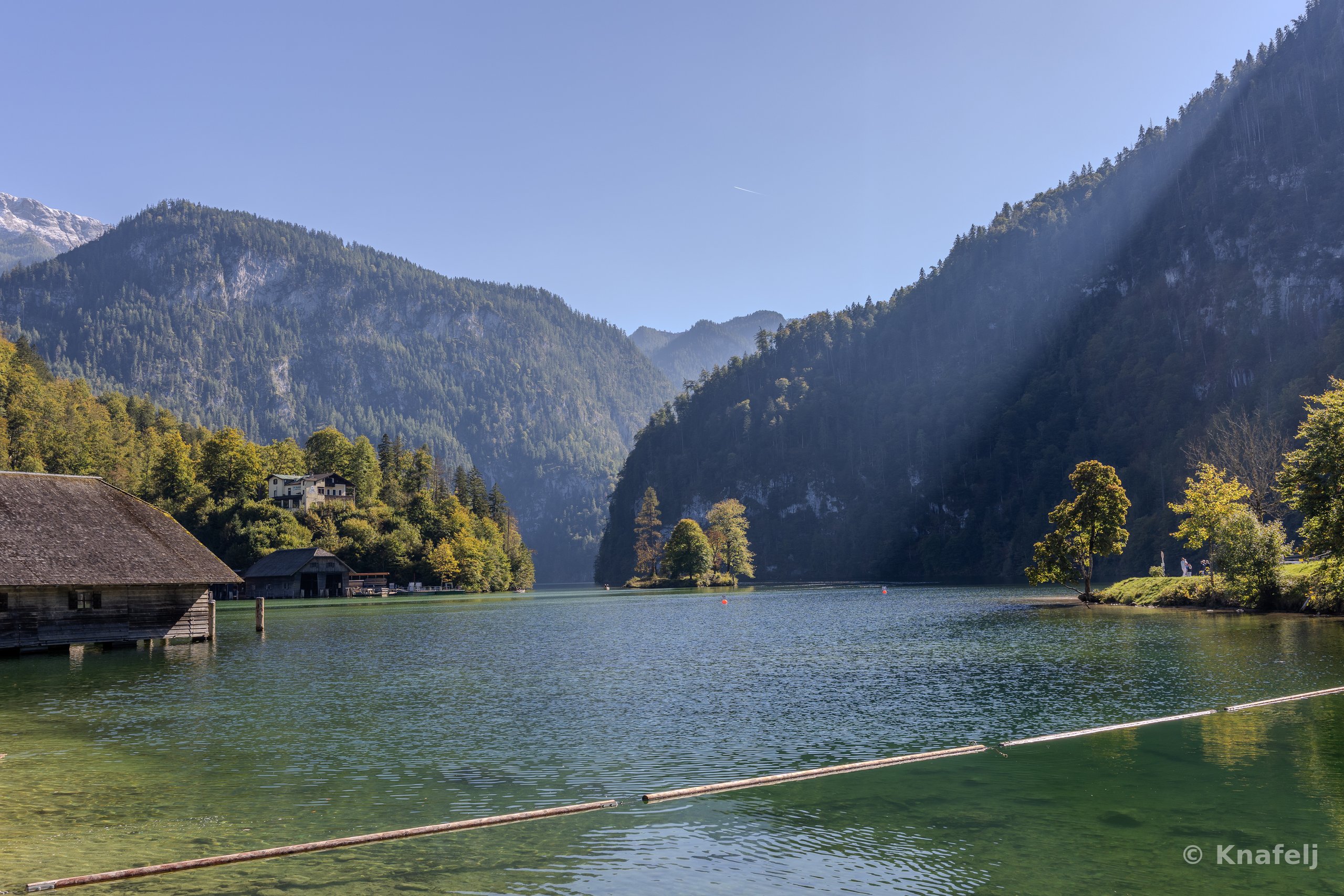 A calm green-blue lake surrounded by steep forested mountains under a clear sky, with old wooden boathouses on the left shore and sunbeams crossing the scene.