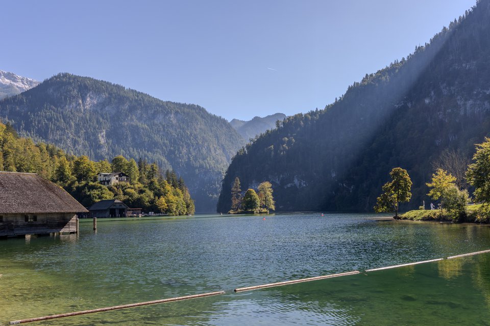 A calm green-blue lake surrounded by steep forested mountains under a clear sky, with old wooden boathouses on the left shore and sunbeams crossing the scene.