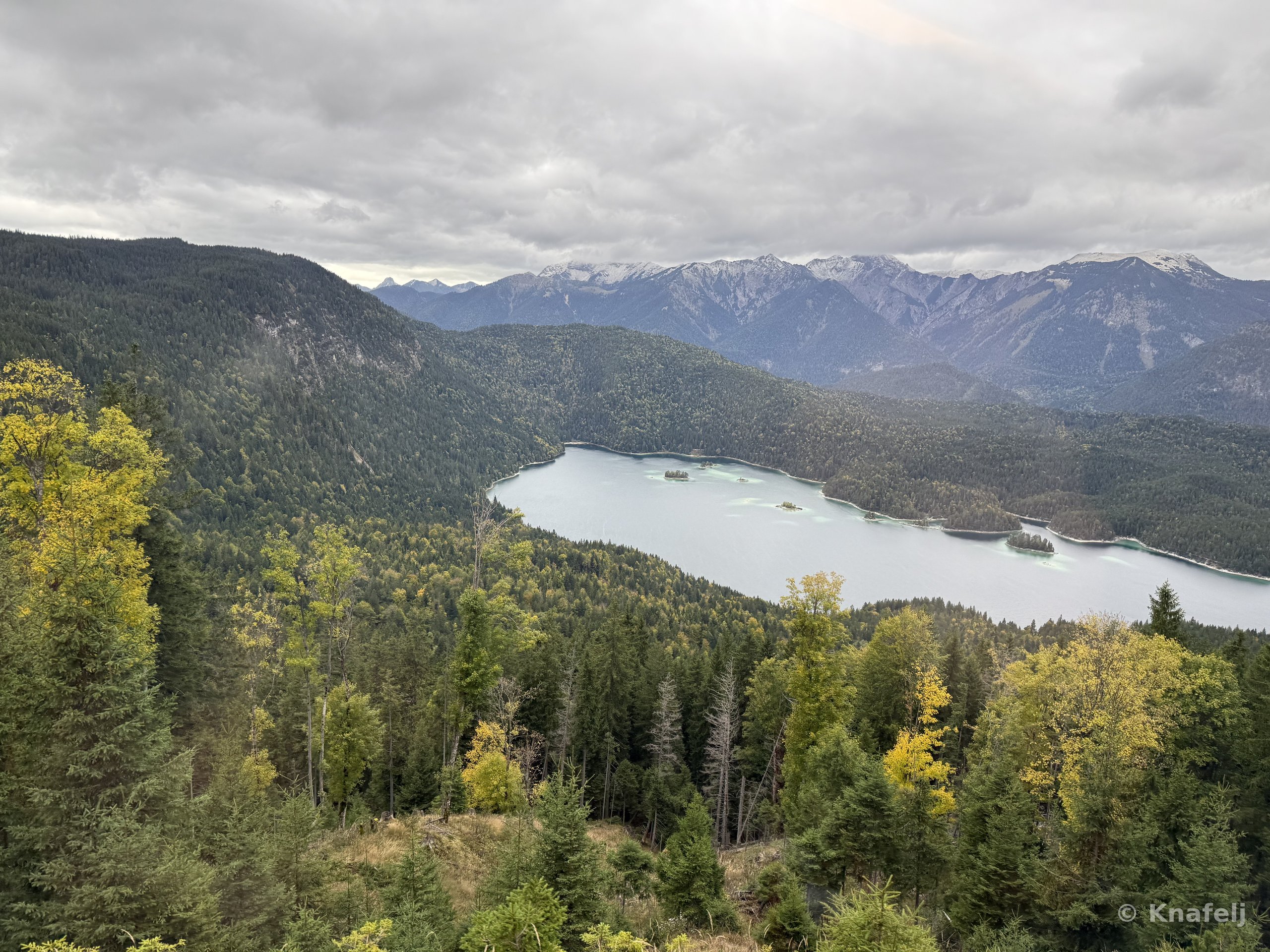 Elevated view of a blue-green lake surrounded by dense evergreen forest, with layered mountain ranges and cloudy skies in the background.