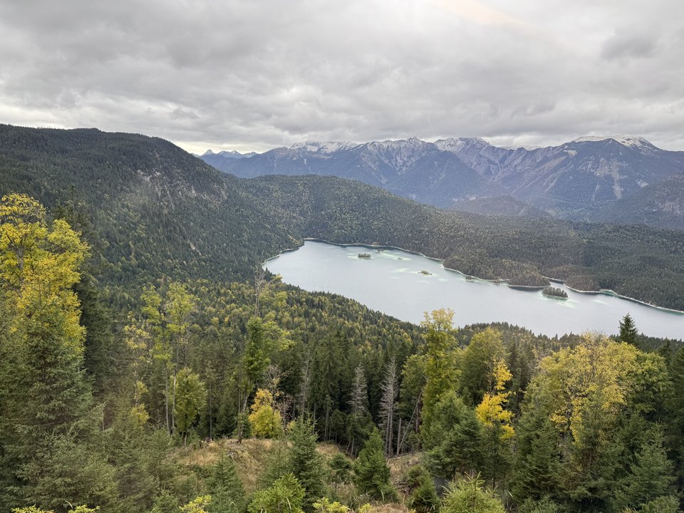 Elevated view of a blue-green lake surrounded by dense evergreen forest, with layered mountain ranges and cloudy skies in the background.