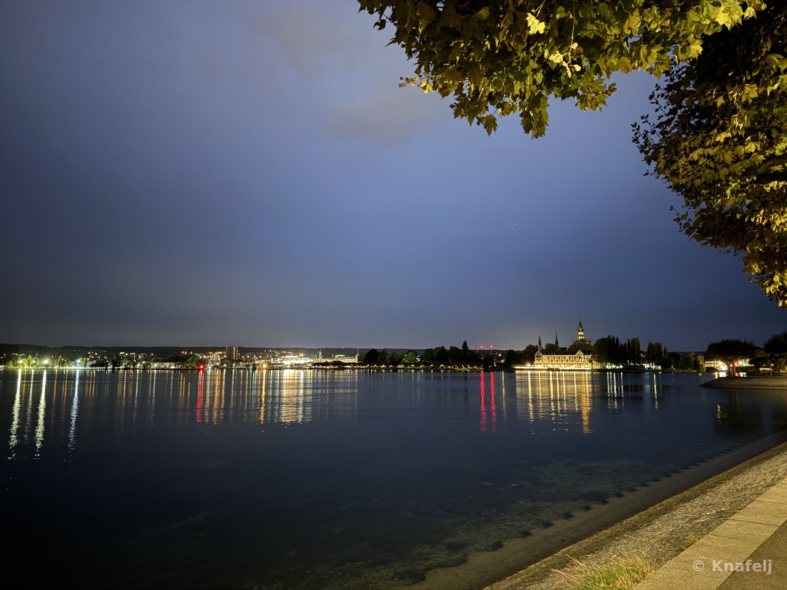 A lakeside at dusk with distant illuminated buildings and a church spire reflected in the still water. The dark blue sky fills the upper half of the frame, and tree leaves hang in the top right corner. A stone walkway is visible in the lower right.