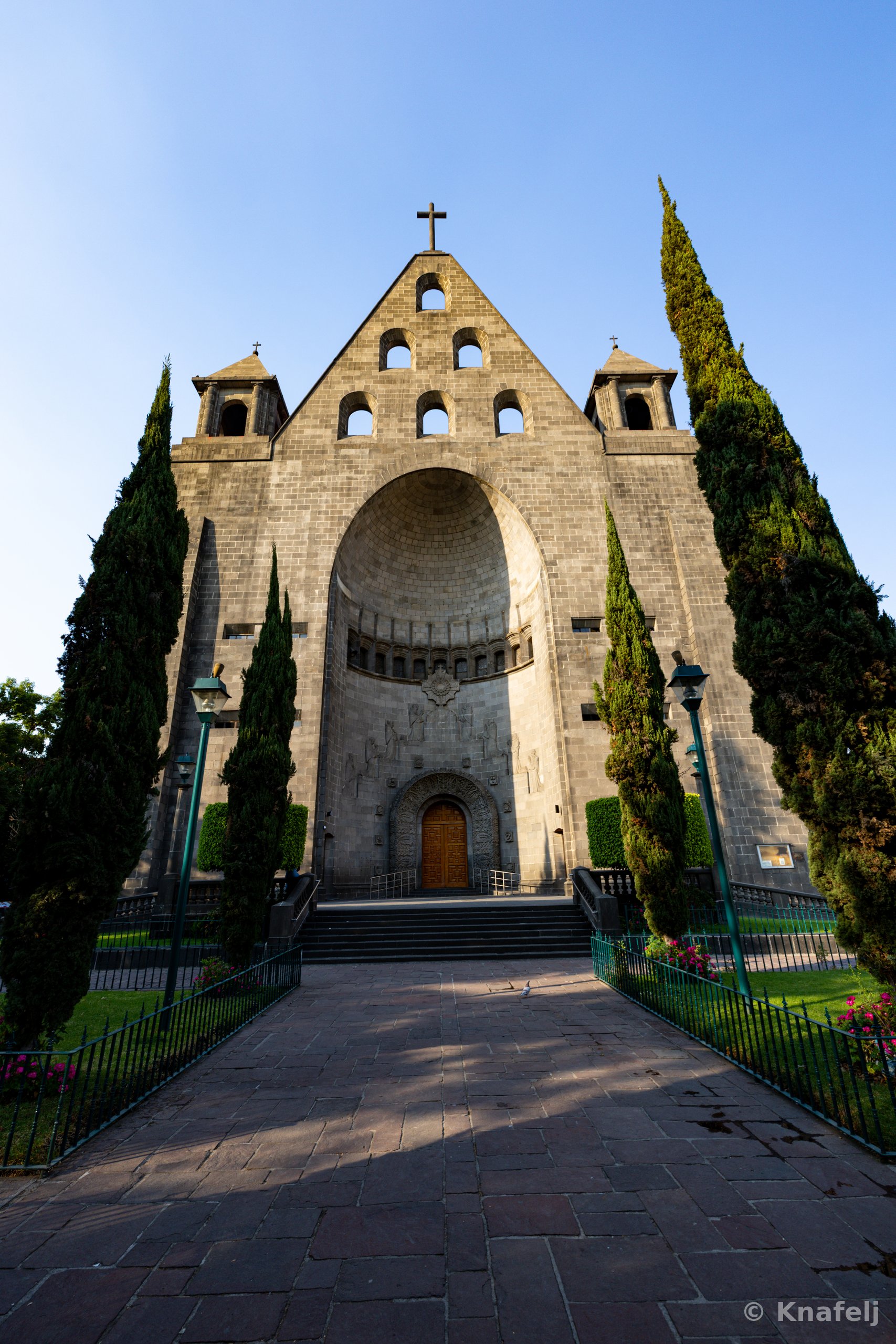 Large gray stone church with a pointed gable, round windows, a cross at the top, and a tall arched entrance, framed by tall cypress trees and approached by a brick pathway under a clear sky.
