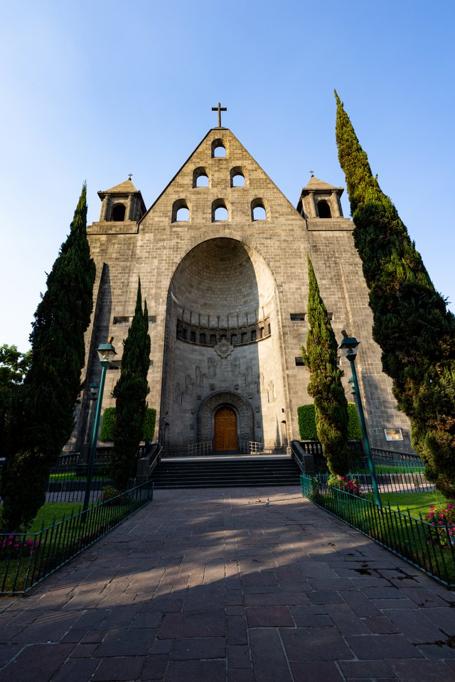 Large gray stone church with a pointed gable, round windows, a cross at the top, and a tall arched entrance, framed by tall cypress trees and approached by a brick pathway under a clear sky.