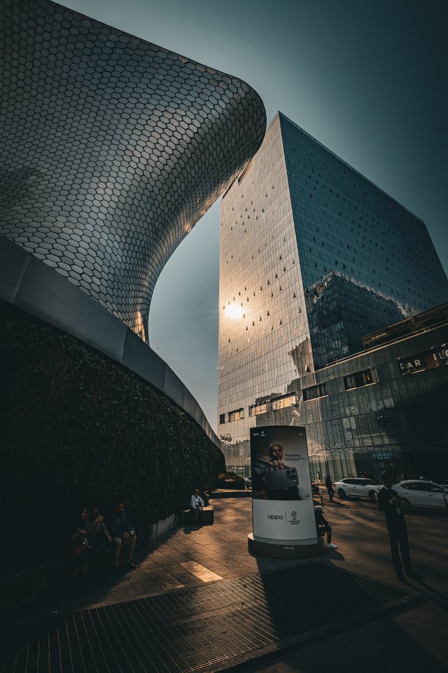 A curving silver hexagon-tiled museum building stands next to a tall glass skyscraper with the sun shining through the gap between them, while people sit on benches and an OPPO advertising column stands in the plaza below.