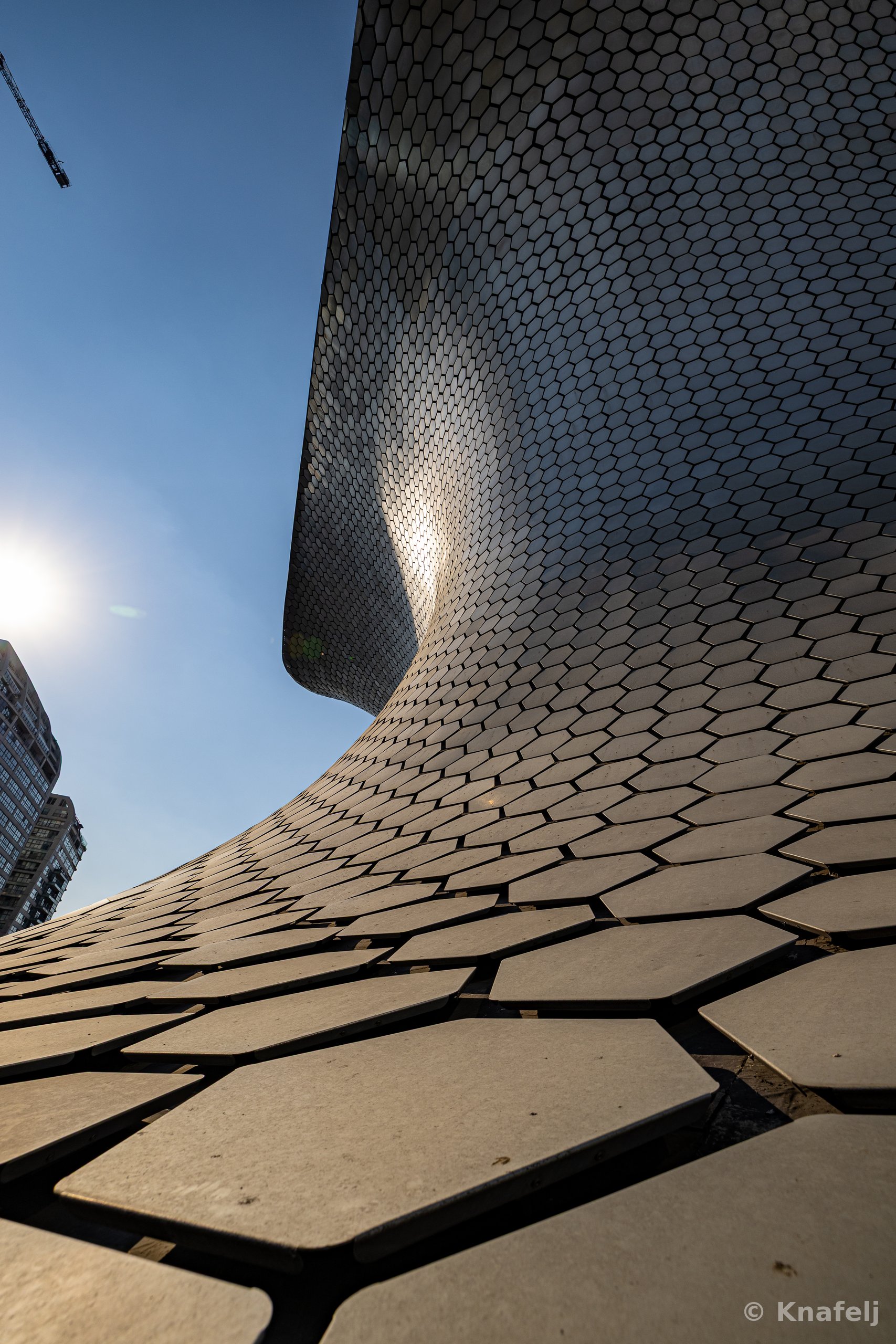 Low-angle view looking up the curved silver hexagonal-tiled exterior of a modern museum building, with the sun flaring on the left, a high-rise tower, and a construction crane against a blue sky.