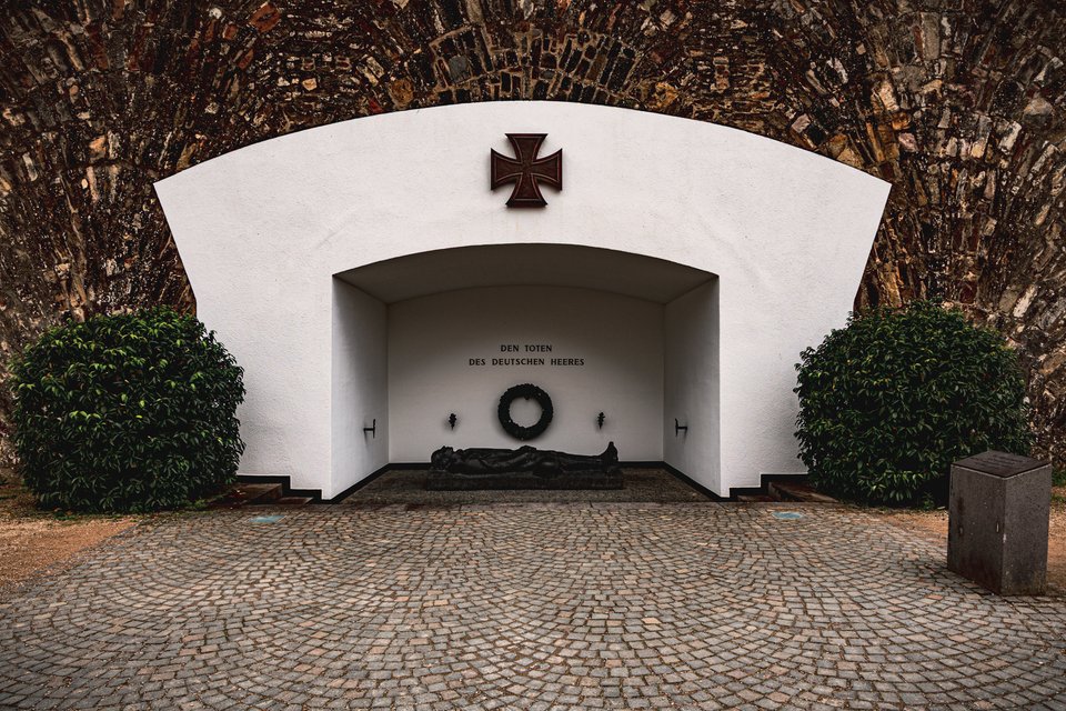 A white memorial structure built into a stone archway, displaying a dark Iron Cross above an inscription reading 'Den Toten des Deutschen Heeres,' with a wreath and a reclining sculpted figure inside, flanked by two rounded green bushes over a cobblestone courtyard.