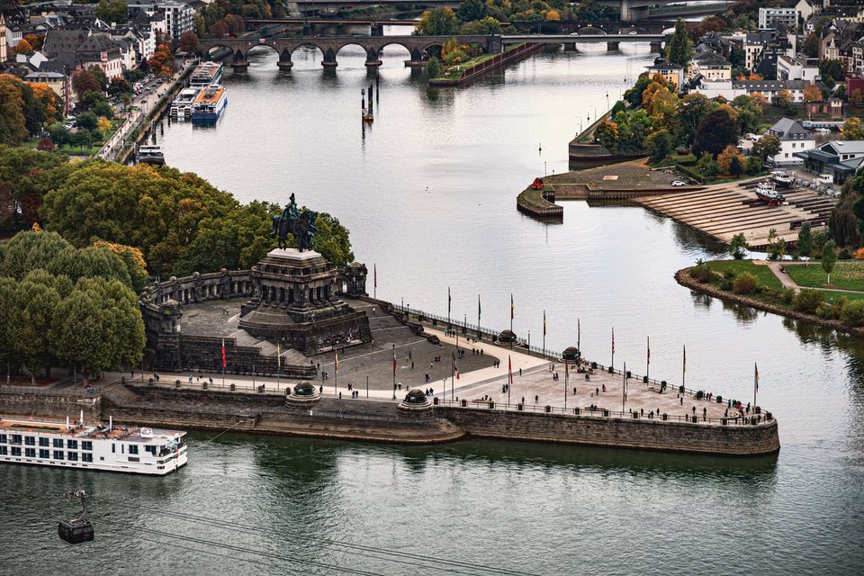 Aerial view of a stone promontory with an equestrian statue monument at the confluence of two rivers, lined with flagpoles and surrounded by river cruise boats, a multi-arched bridge, and a city in the background.