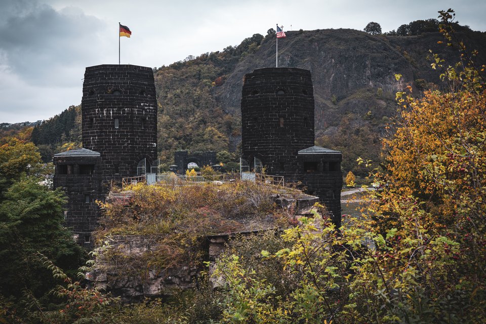 Two tall dark stone towers with conical bases stand before a forested hill under a cloudy sky. The left tower flies a German flag and the right flies an American flag. Autumn-colored bushes and trees grow in the foreground around the ruins.
