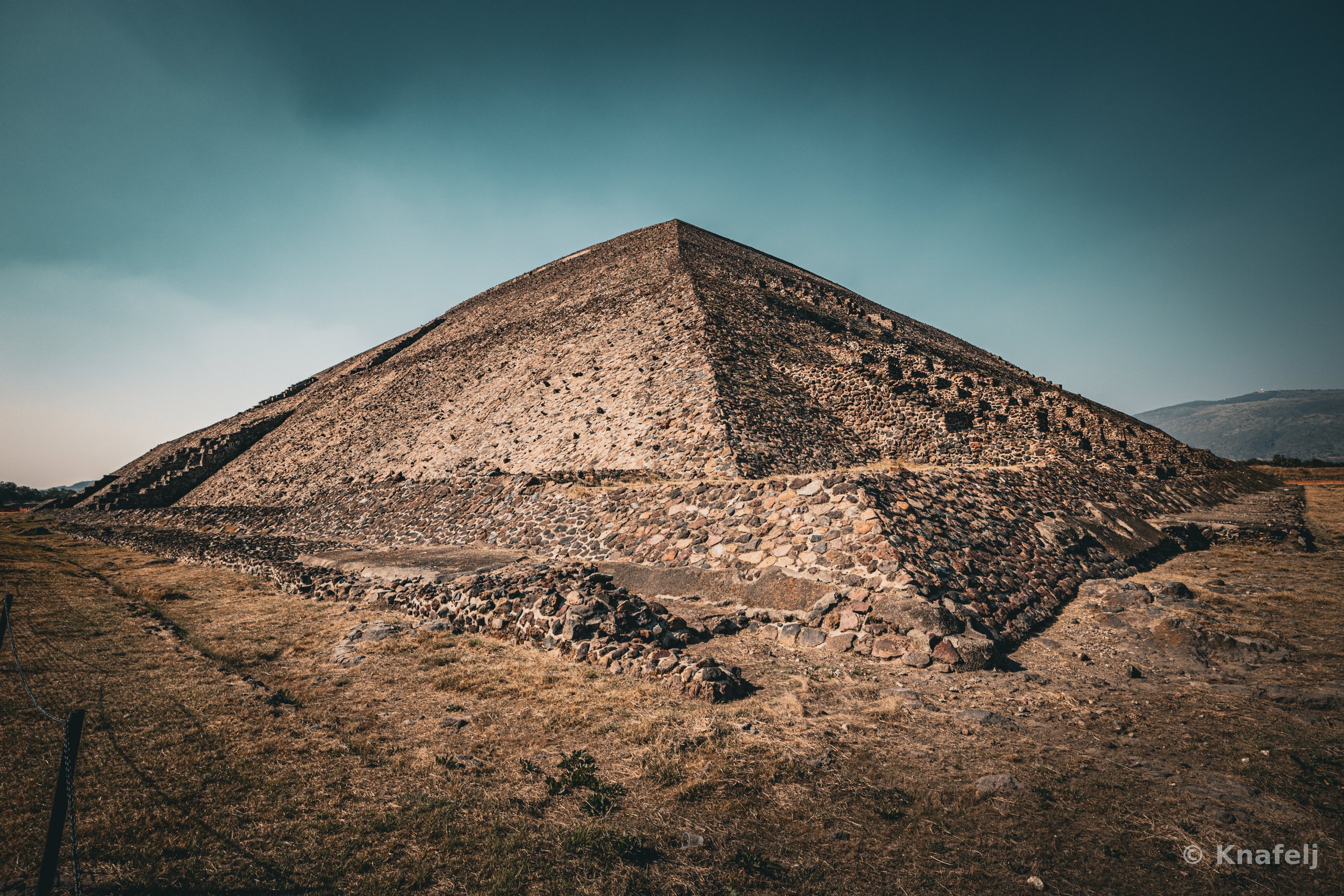 A large stepped stone pyramid photographed from its corner, set against a dark teal and gray sky, with dry brown grass in the foreground and distant hills to the right.