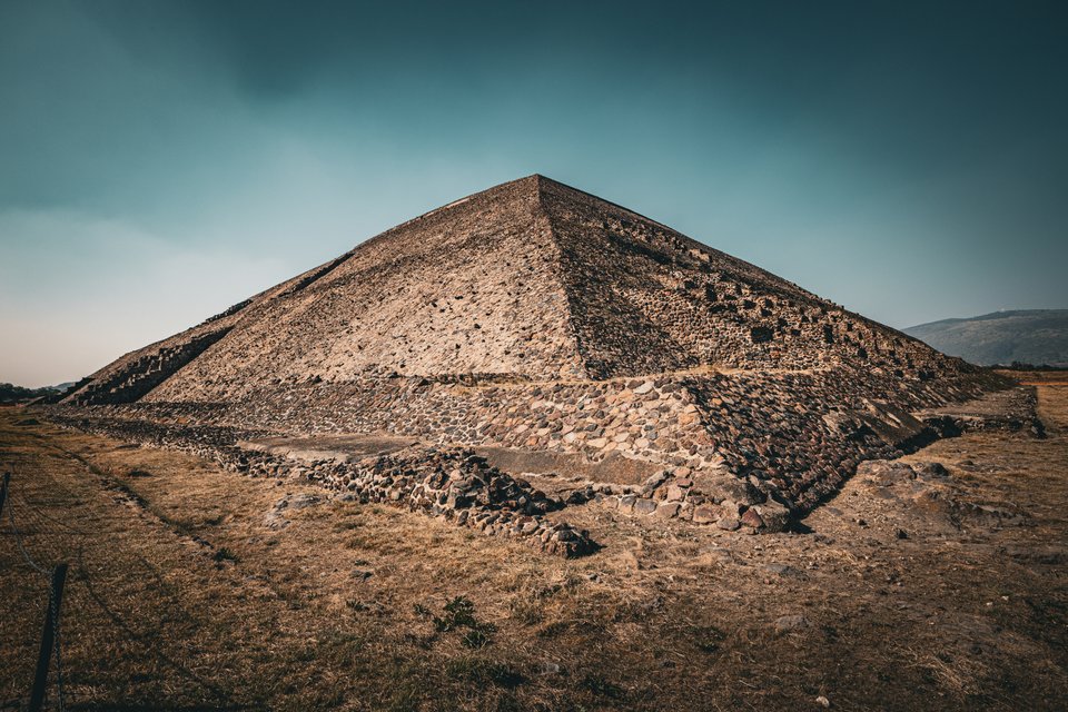 A large stepped stone pyramid photographed from its corner, set against a dark teal and gray sky, with dry brown grass in the foreground and distant hills to the right.