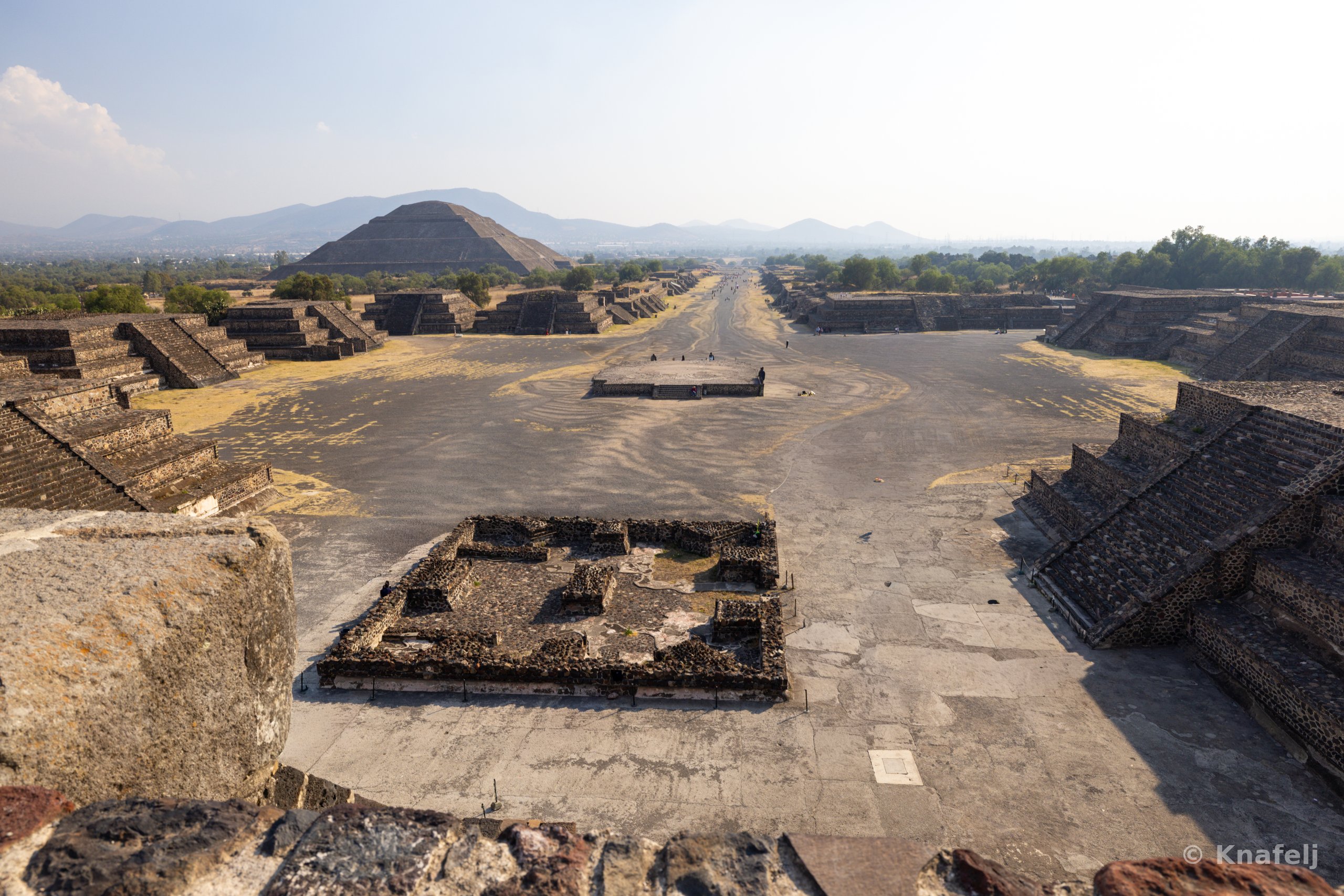 Aerial view from atop a pyramid looking over the ruins of Teotihuacan, showing a long central avenue flanked by stone platforms and structures, with a large pyramid on the left and distant mountains under a pale sky.