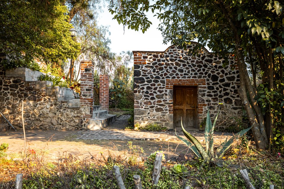An old stone house built with rounded volcanic stones and red brick accents, featuring a wooden door, set in a green garden with trees, an agave plant in the foreground, and a cobblestone walkway.