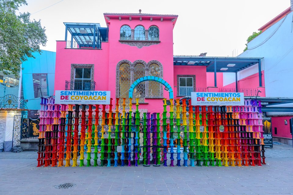 A colorful outdoor art installation of vertical columns in rainbow hues arranged in front of a pink two-story colonial building with arched windows and signage reading 'Sentimientos de Coyoacan'.