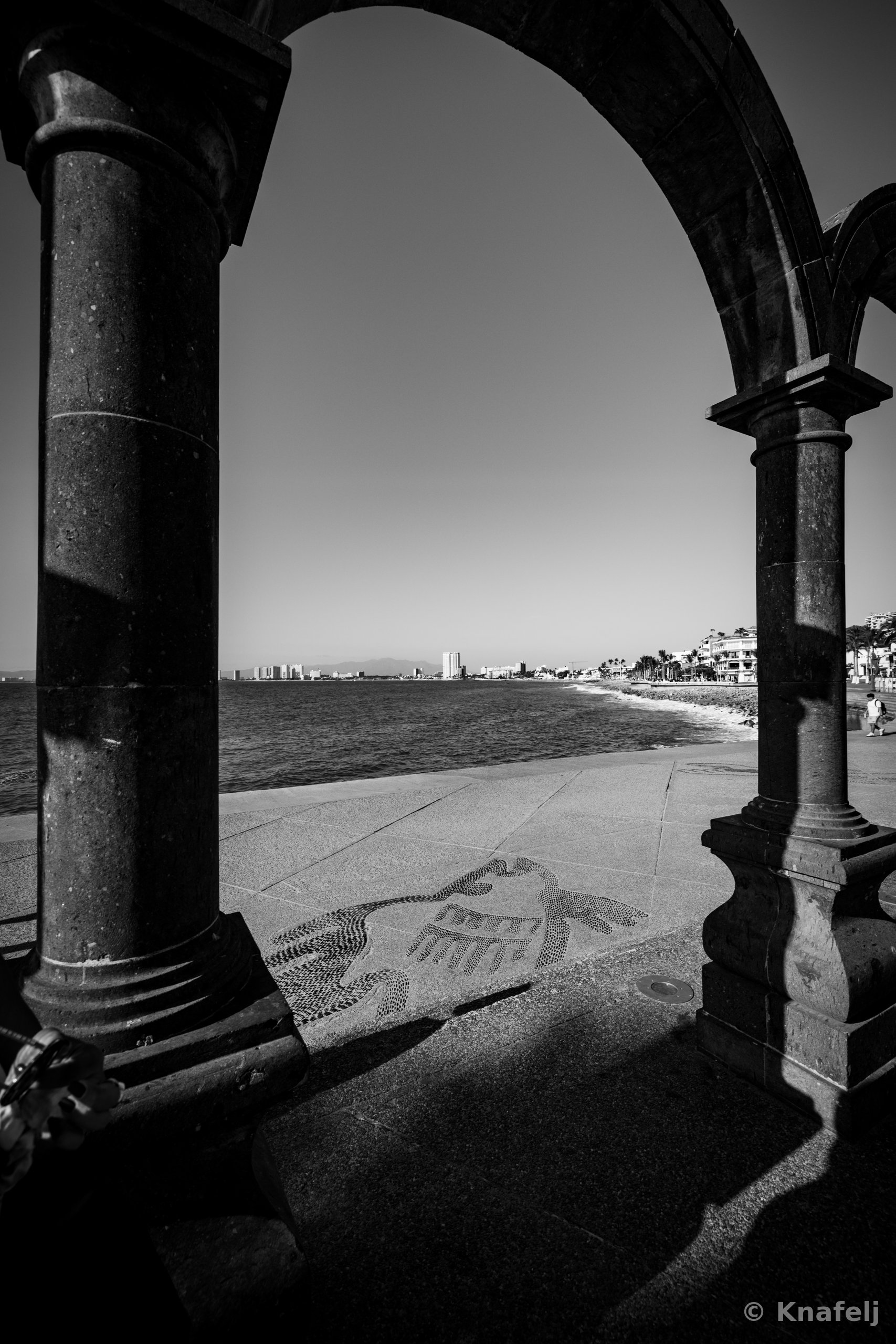 Black and white photo looking through a large stone arch and columns onto a seaside walkway with a decorative tile mosaic on the pavement, calm water, and a distant skyline under a clear sky.