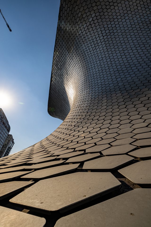 Low-angle view looking up the curved silver hexagonal-tiled exterior of a modern museum building, with the sun flaring on the left, a high-rise tower, and a construction crane against a blue sky.