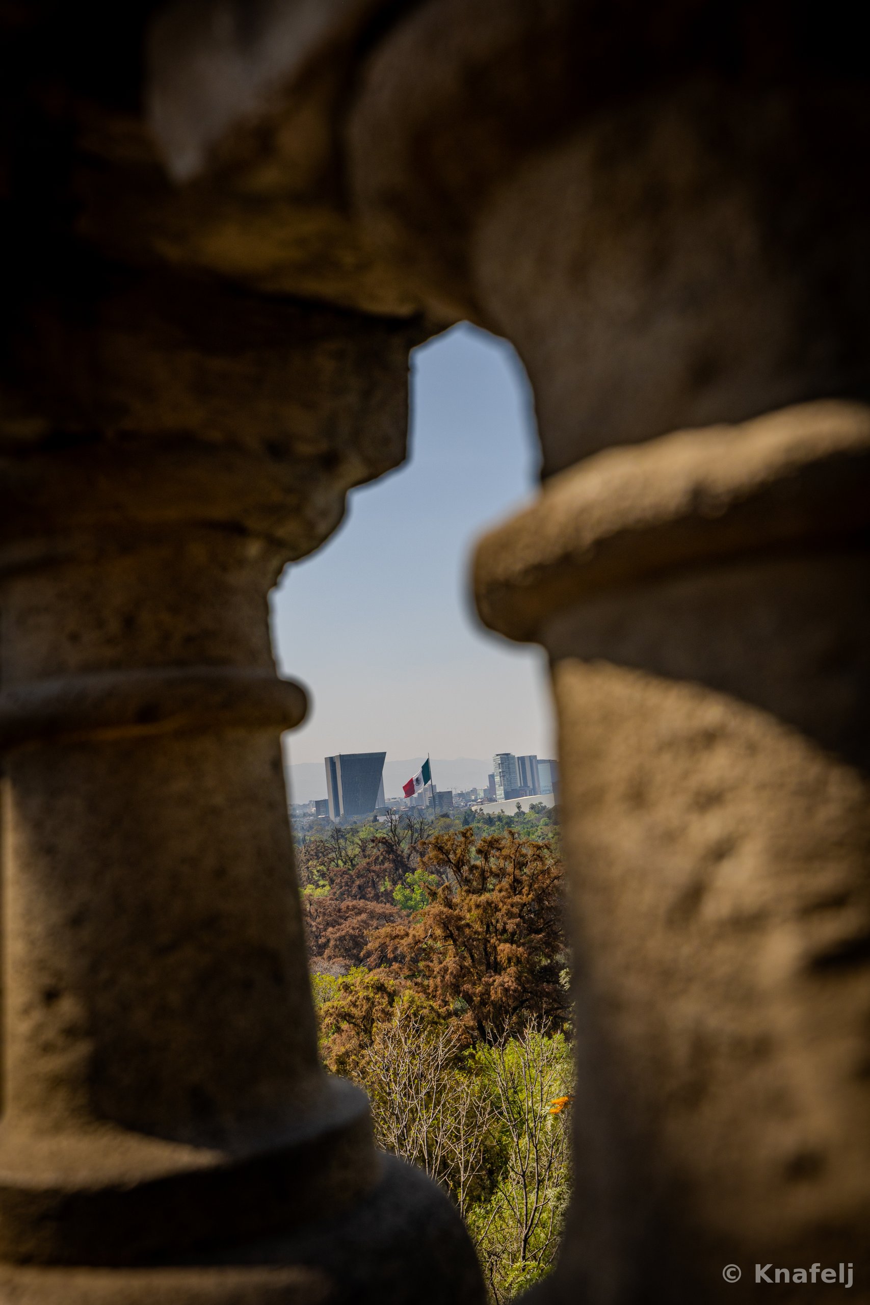 View through two stone balustrade columns showing a hazy city skyline with a Mexican flag in the distance, above a field of green and brown treetops under a pale blue sky.