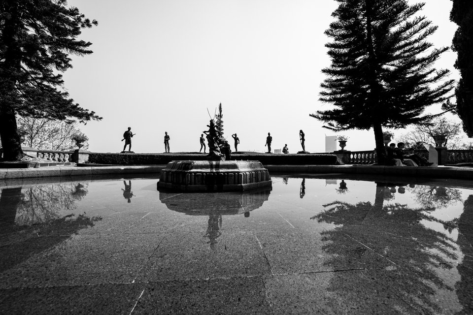 Black and white photo of a garden terrace with a stone fountain and silhouetted human figures and statues lined up against a pale sky, framed by tall pine trees, with reflections cast in a still pool in the foreground.