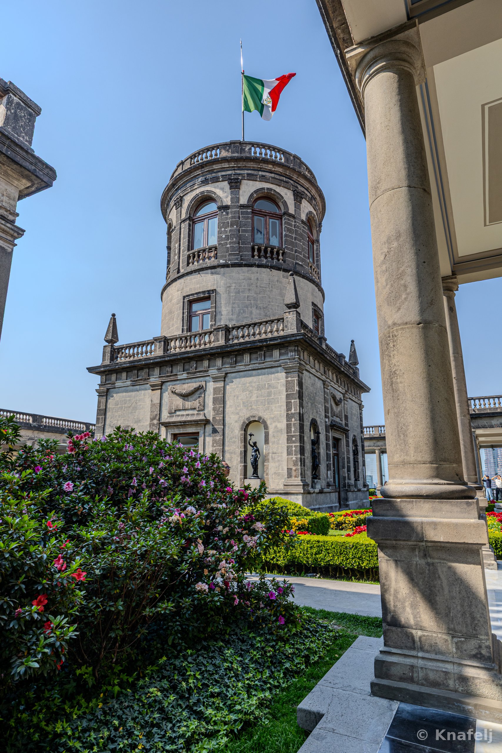 A circular stone tower of a historic castle topped with the Mexican flag, viewed from a garden with blooming flowers, hedges, and a large stone column in the foreground under a clear sky.