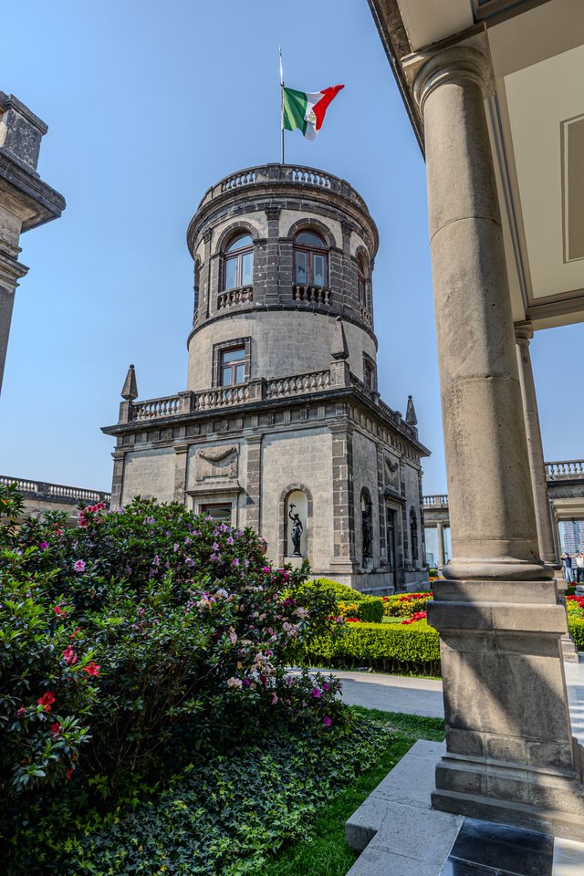 A circular stone tower of a historic castle topped with the Mexican flag, viewed from a garden with blooming flowers, hedges, and a large stone column in the foreground under a clear sky.