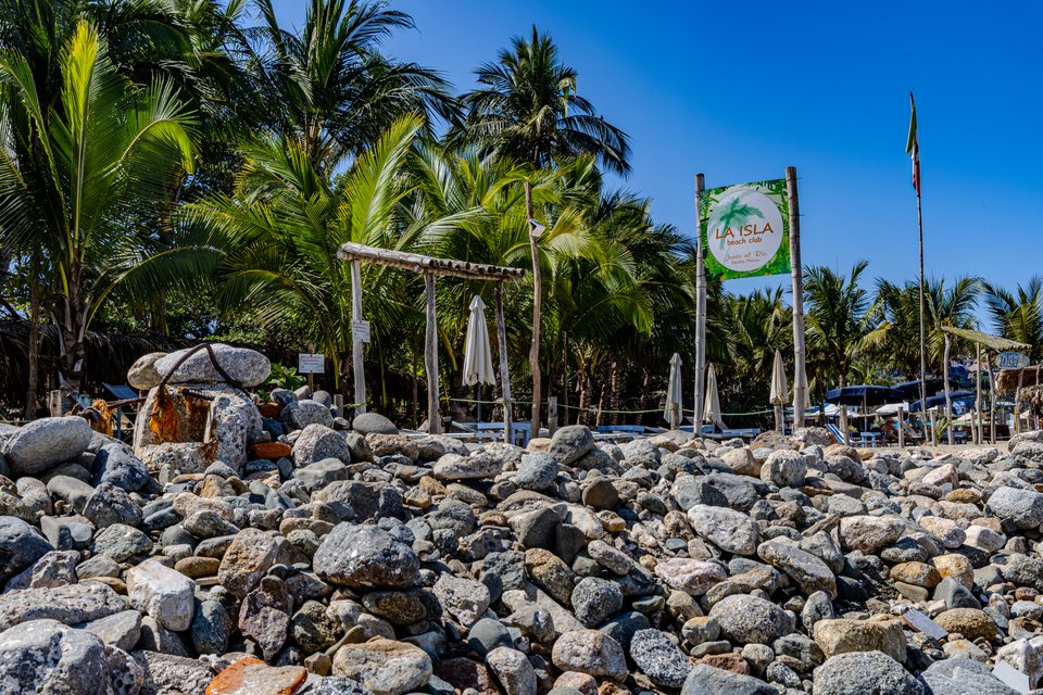 A pebble and stone-covered beach with a wooden archway and a sign reading La Isla Beach Club, surrounded by tall palm trees and folded white umbrellas against a clear blue sky.