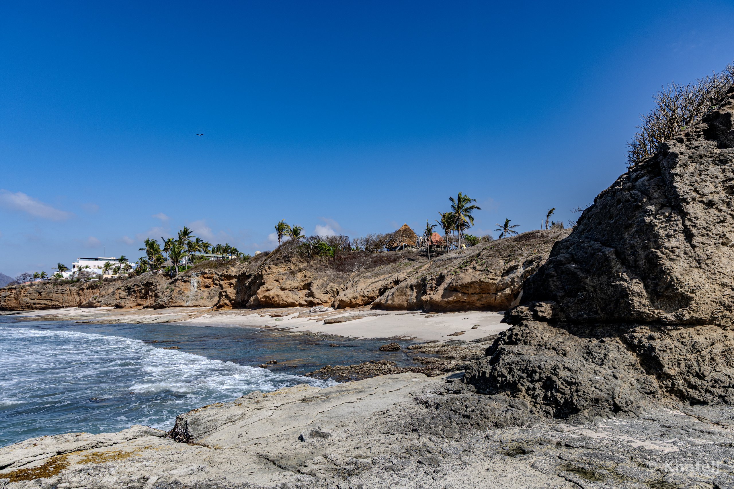 A coastal scene with a large rocky outcrop on the right, a small sandy beach with gentle waves on the left, palm trees and buildings on a cliff in the background, under a deep blue sky.