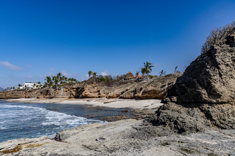A coastal scene with a large rocky outcrop on the right, a small sandy beach with gentle waves on the left, palm trees and buildings on a cliff in the background, under a deep blue sky.