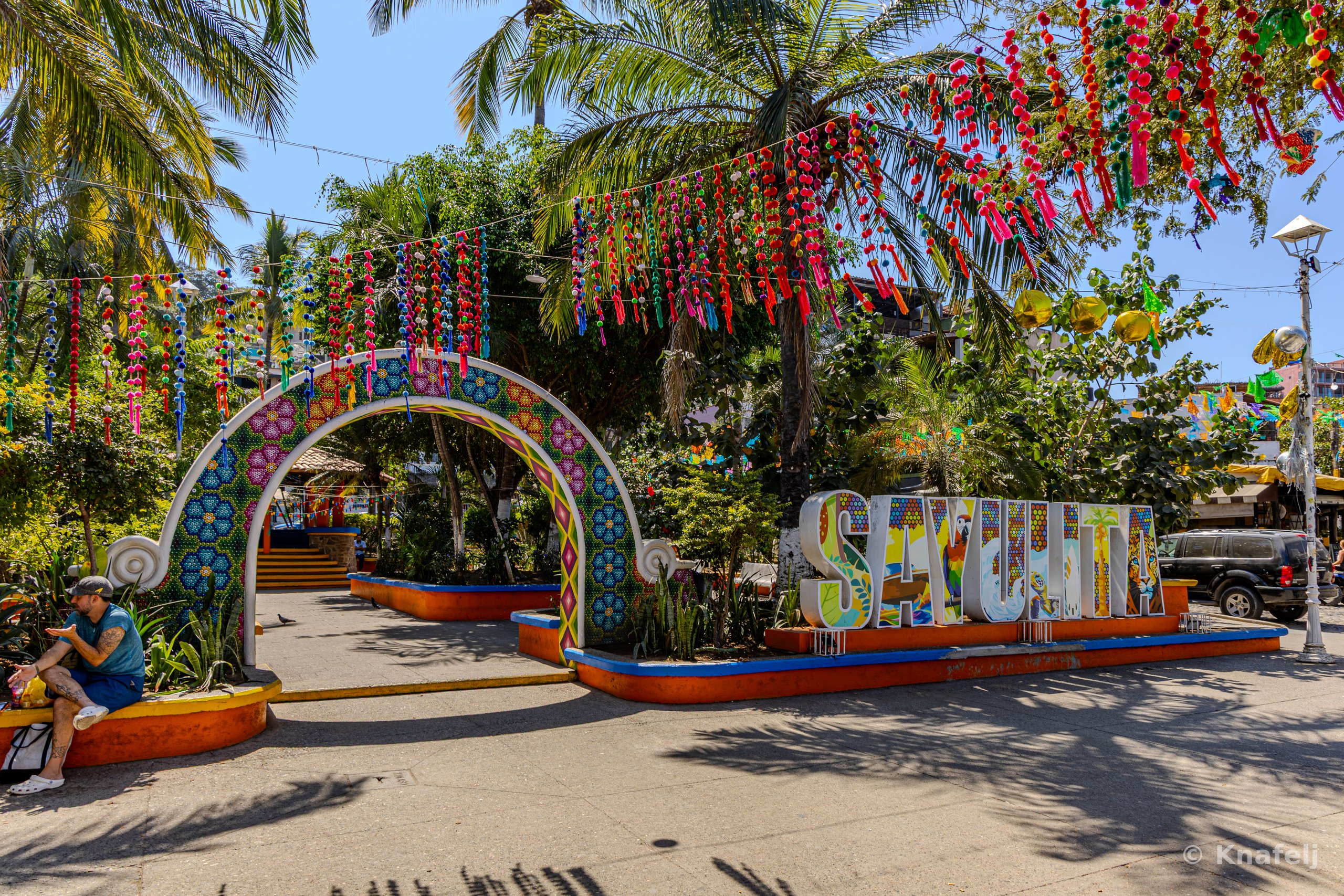 A decorated town square with a colorful mosaic-tiled arch, multicolored ribbon decorations strung overhead, large painted letters spelling SAYULITA, palm trees, and a man sitting on a ledge to the left.