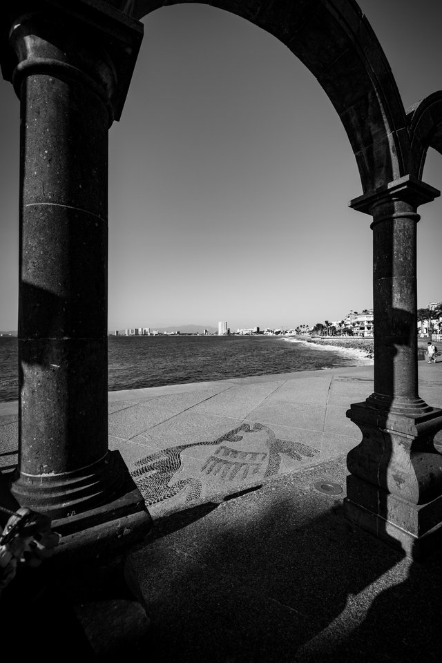 Black and white photo looking through a large stone arch and columns onto a seaside walkway with a decorative tile mosaic on the pavement, calm water, and a distant skyline under a clear sky.