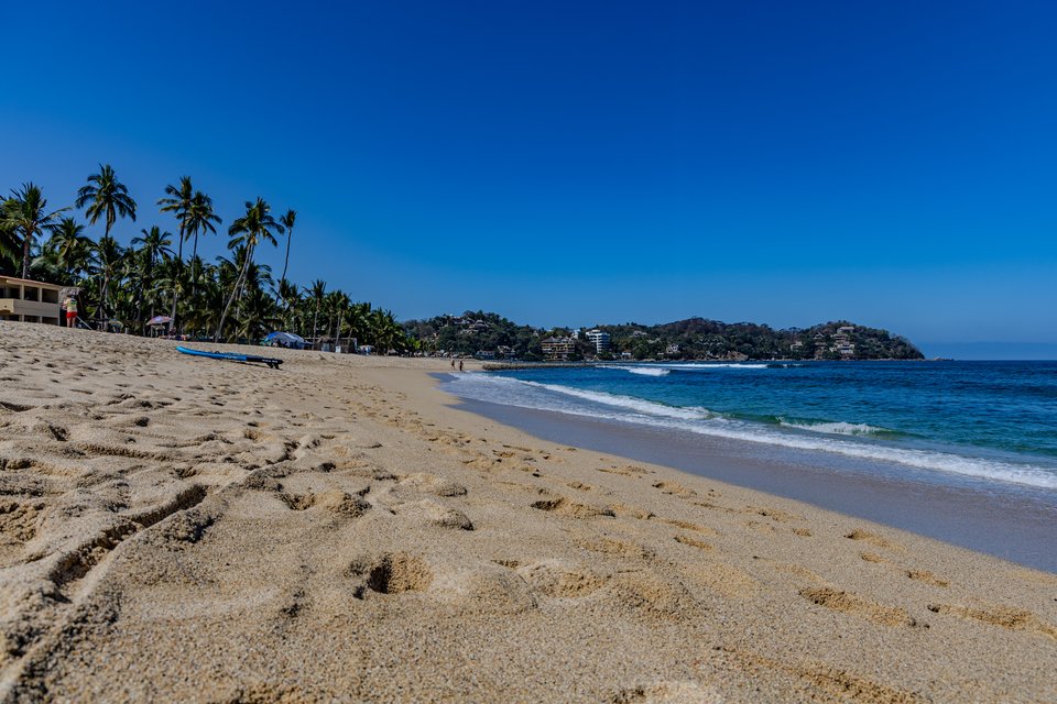 Wide beach with golden sand in the foreground, palm trees along the left shoreline, turquoise ocean waves breaking on the right, and forested hills under a deep blue sky.
