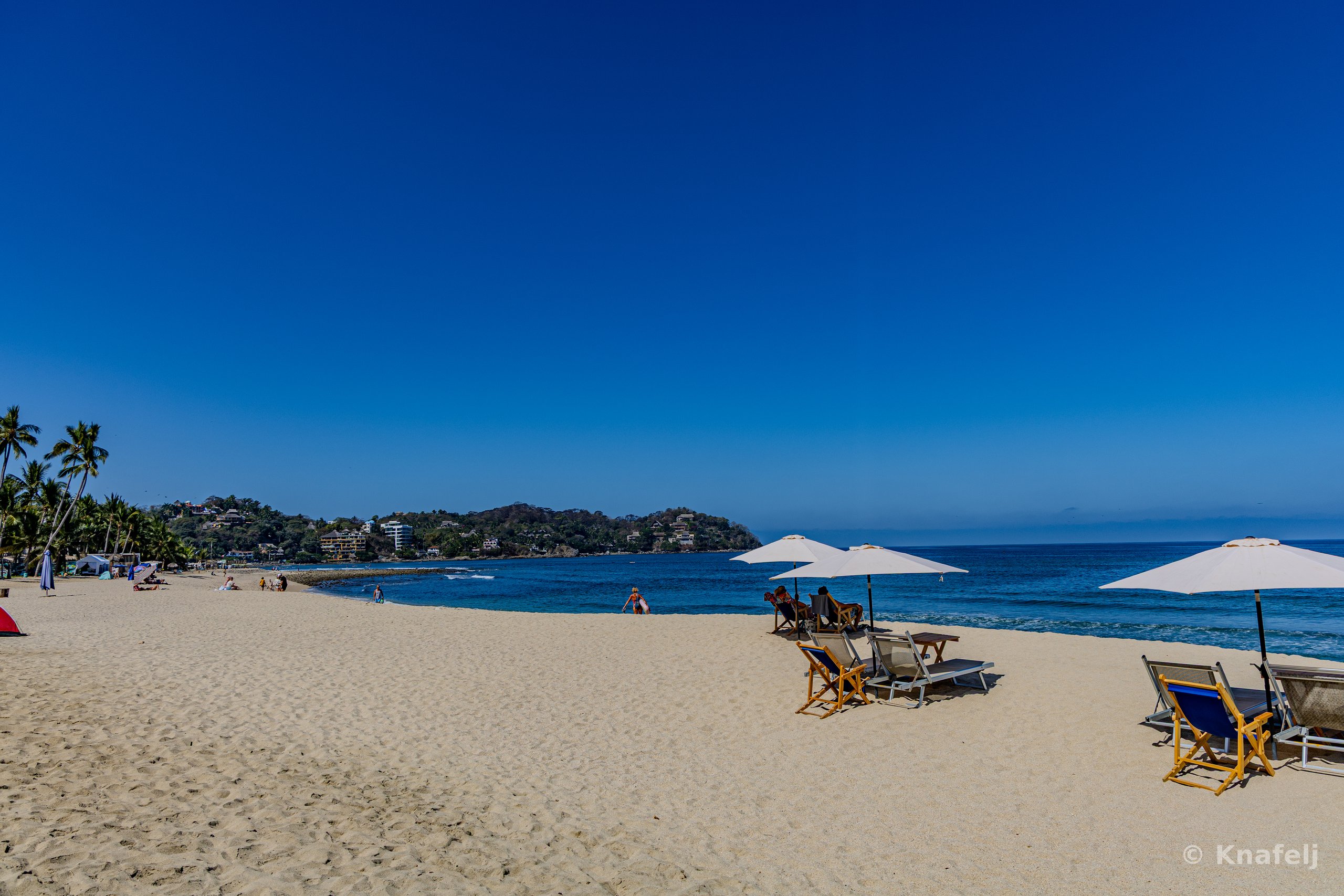 A wide sandy beach with white sun umbrellas and lounge chairs in the foreground, a calm blue ocean bay, distant green hills, and palm trees on the left under a clear blue sky.