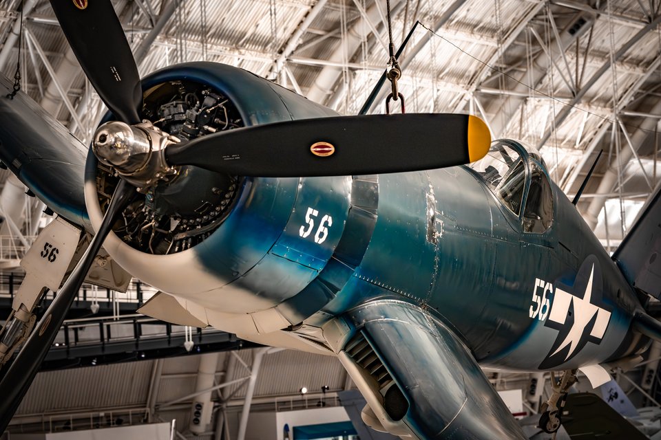 A blue WWII naval fighter aircraft with number 56 and a white star insignia suspended from the ceiling of a museum, showing its radial engine, propeller, and cockpit beneath exposed steel roof trusses.