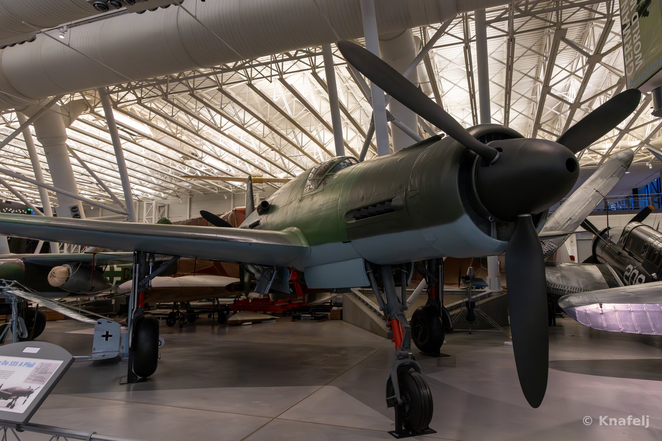 A green and gray German World War II fighter aircraft with a large black propeller nose, parked indoors in a museum hangar with a lattice roof and other vintage planes in the background.
