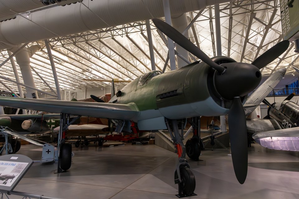 A green and gray German World War II fighter aircraft with a large black propeller nose, parked indoors in a museum hangar with a lattice roof and other vintage planes in the background.