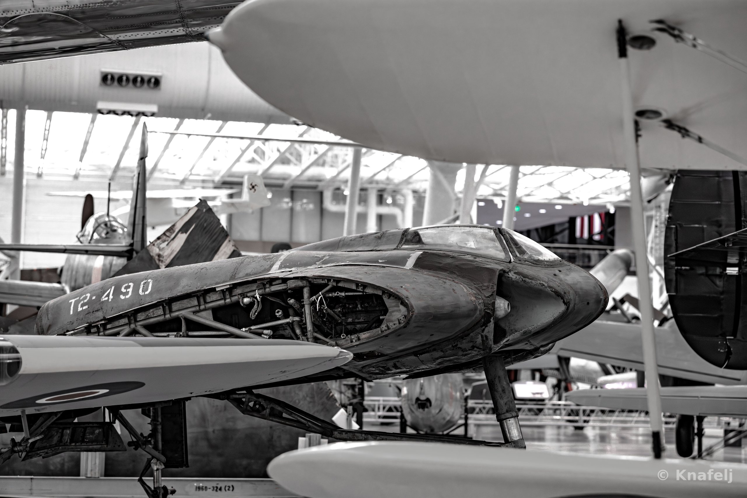 A partially disassembled experimental aircraft fuselage displayed indoors among other vintage planes in a large museum hangar, shown in near black and white with visible internal structure.