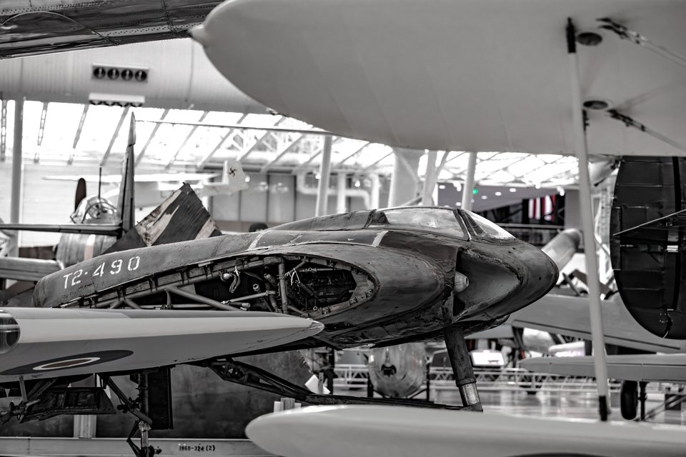 A partially disassembled experimental aircraft fuselage displayed indoors among other vintage planes in a large museum hangar, shown in near black and white with visible internal structure.