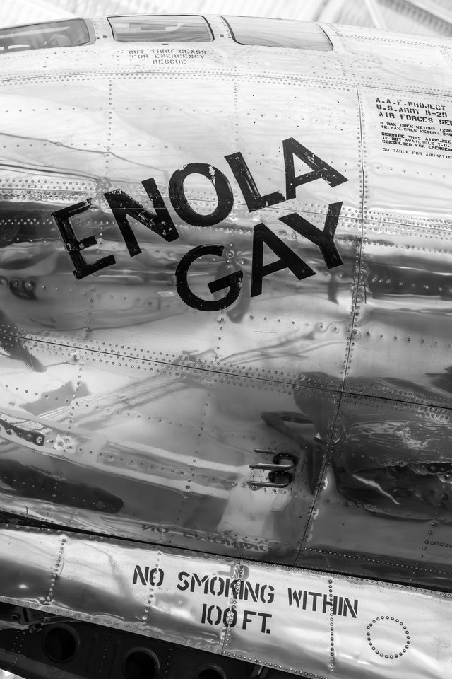 Black and white close-up of a shiny riveted aircraft fuselage with the words 'Enola Gay' painted in large black letters and 'No Smoking Within 100 Ft.' stenciled below.