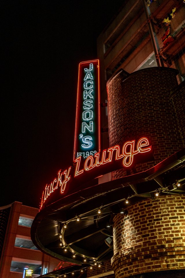 Illuminated vertical neon sign reading 'Jackson's Lucky Lounge' in blue and red letters mounted on a brick building at night, with string lights along the curved facade below.