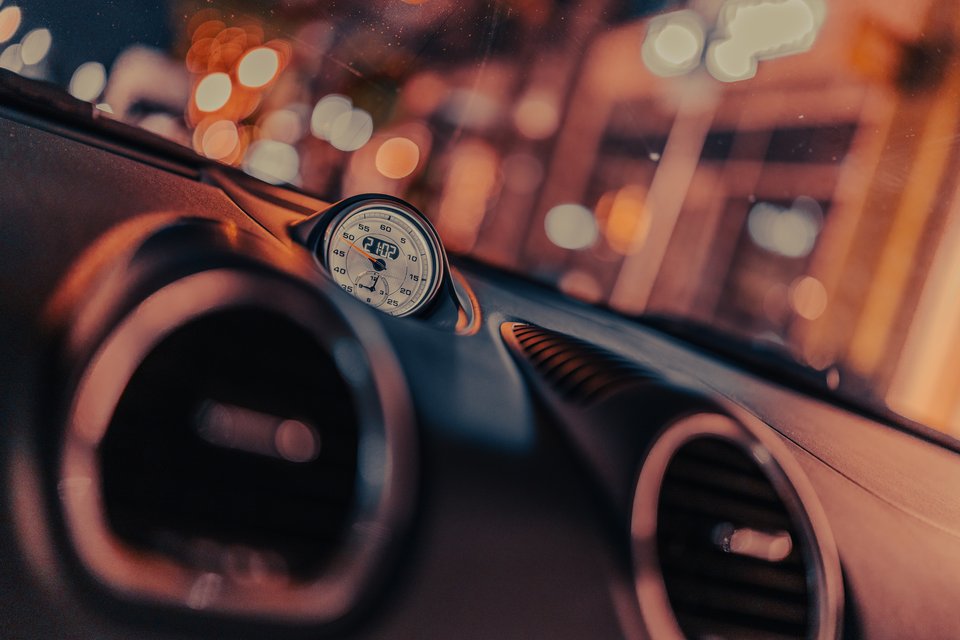 Close-up of a round analog timer gauge on a dark car dashboard, with circular air vents and out-of-focus orange city lights visible through the windshield.