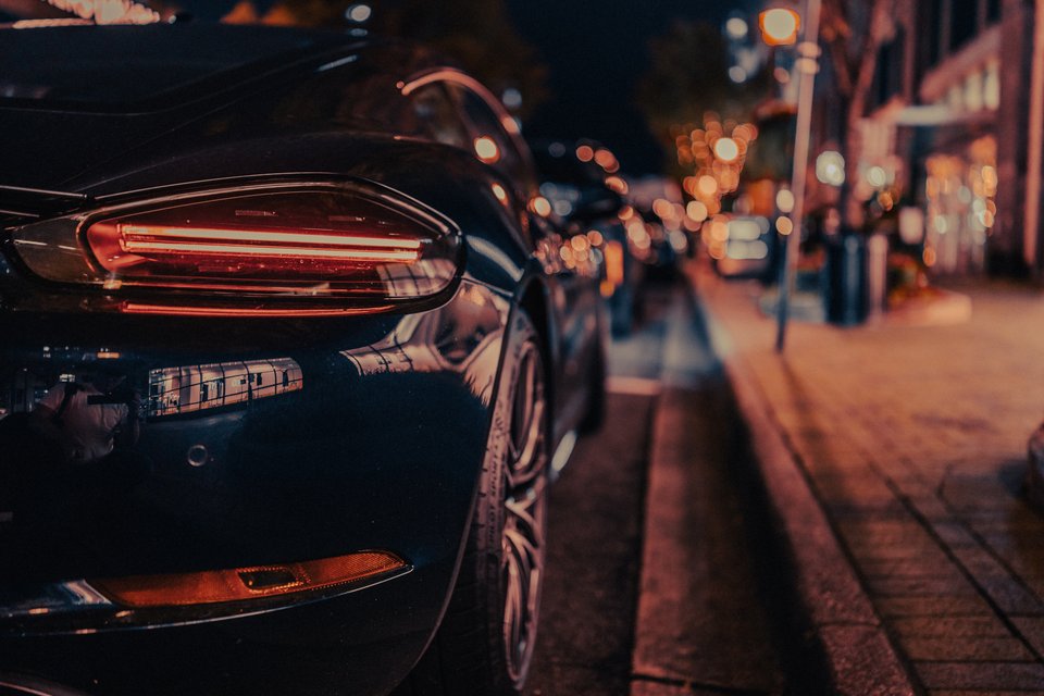 Close-up of the rear taillight and bumper of a dark sports car parked on a city street at night, with blurred warm lights and a sidewalk in the background.