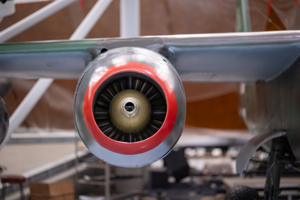 Front view of an aircraft jet engine with a red-painted intake ring and visible turbine fan blades, mounted under a metallic wing in an indoor display.