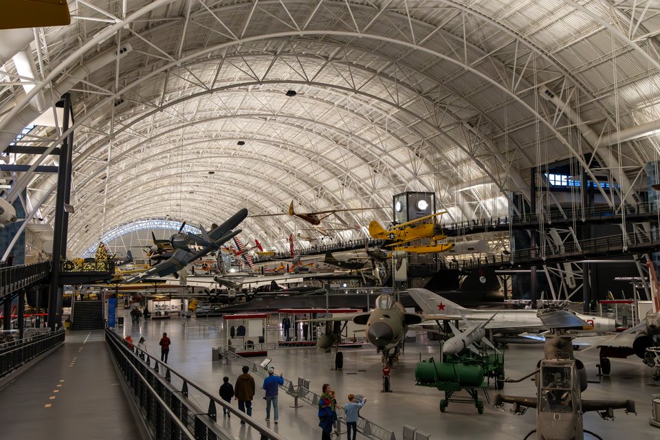Interior of a large aviation museum with a curved metal lattice ceiling. Numerous airplanes are suspended in the air and displayed on the floor, including a yellow biplane and military jets. People walk along an elevated walkway in the foreground.