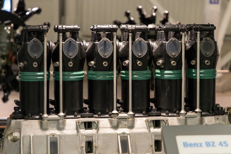 Close-up of a vintage Benz BZ 4 inline engine with six black cylinders, green bands, metallic valve covers, and pushrods, mounted on an aluminum crankcase with a nameplate reading 'Benz BZ 4S'.