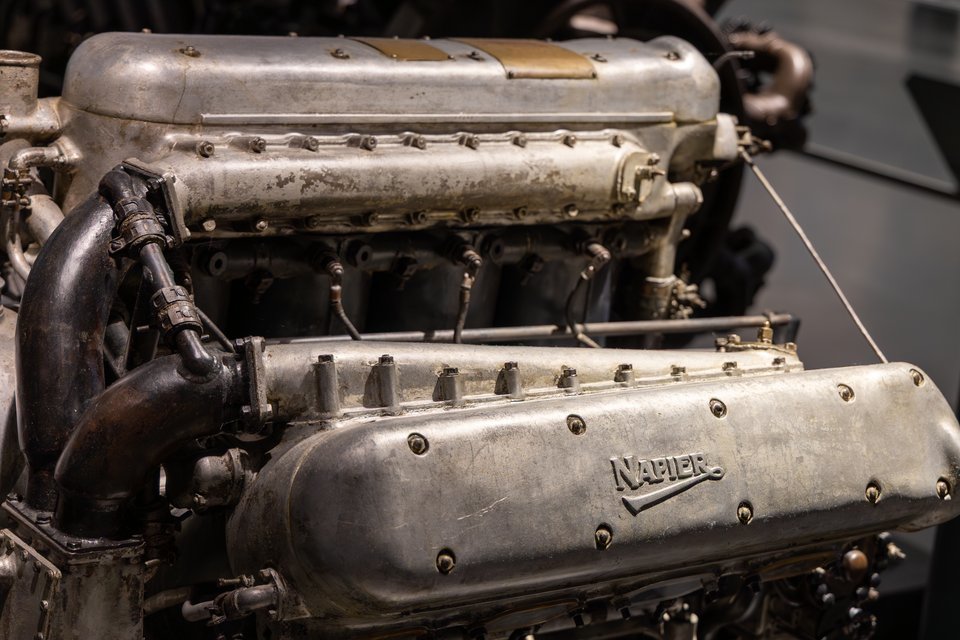 Close-up of a vintage Napier engine with polished and tarnished aluminum cylinder banks, rows of bolts, and dark curved exhaust pipes, embossed with the Napier logo.