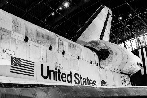 Black and white photograph of the Space Shuttle Discovery's fuselage showing the American flag, 'United States' lettering, and NASA logo, with its tail and tiled surface under a museum's geometric ceiling.