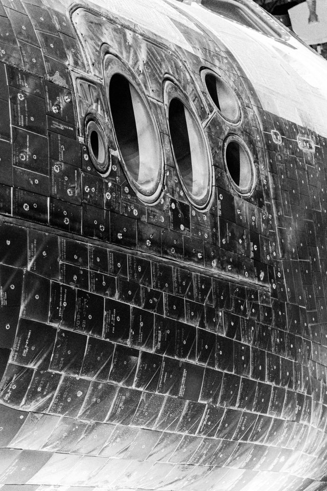 Black and white close-up of a space shuttle's underside and cockpit windows, covered in numbered thermal protection tiles arranged in rows.