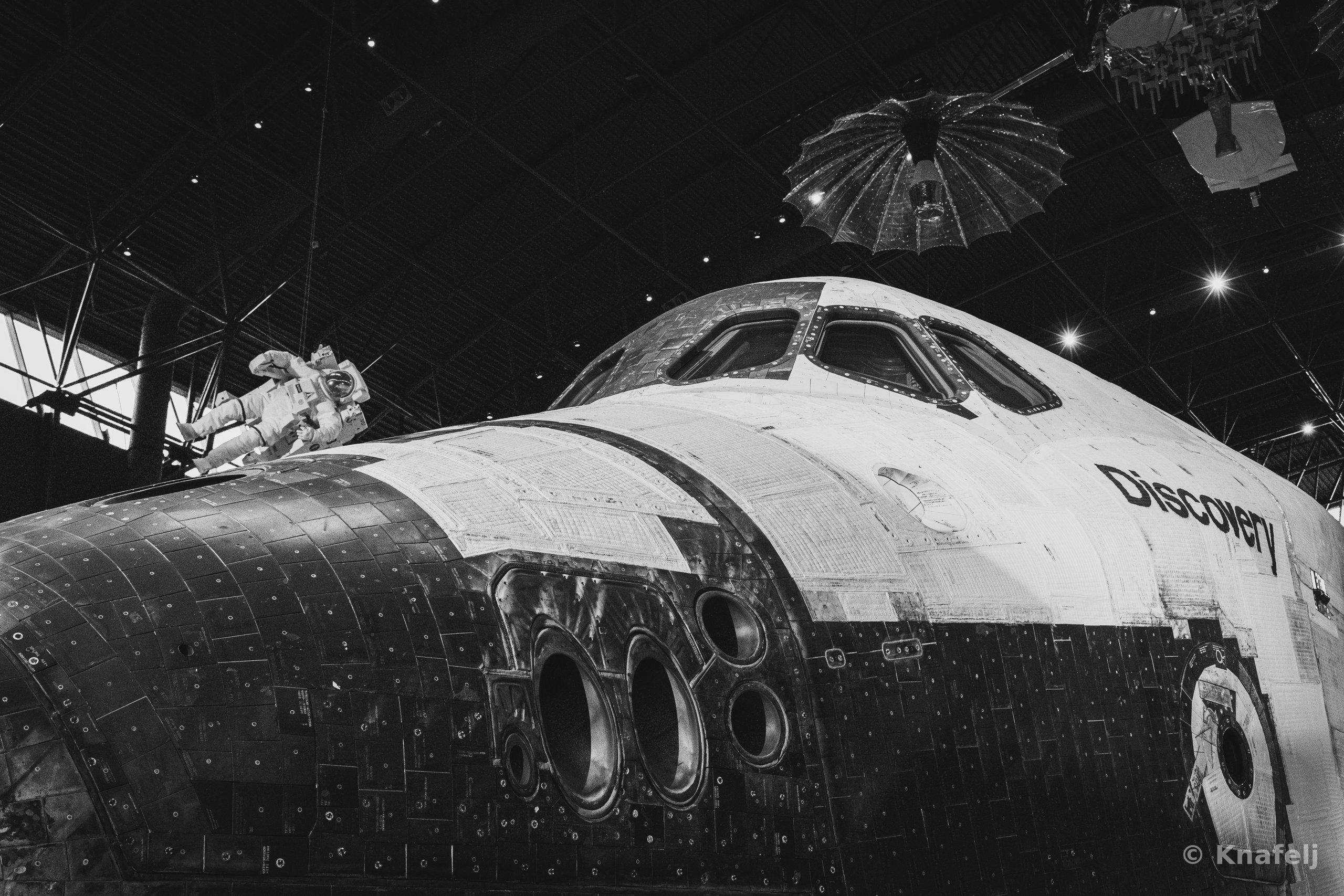 Black and white photo of the Space Shuttle Discovery displayed in a museum, showing its nose, cockpit windows, and tiled underside, with a suspended astronaut figure and parachute-like satellite hanging from the ceiling above.