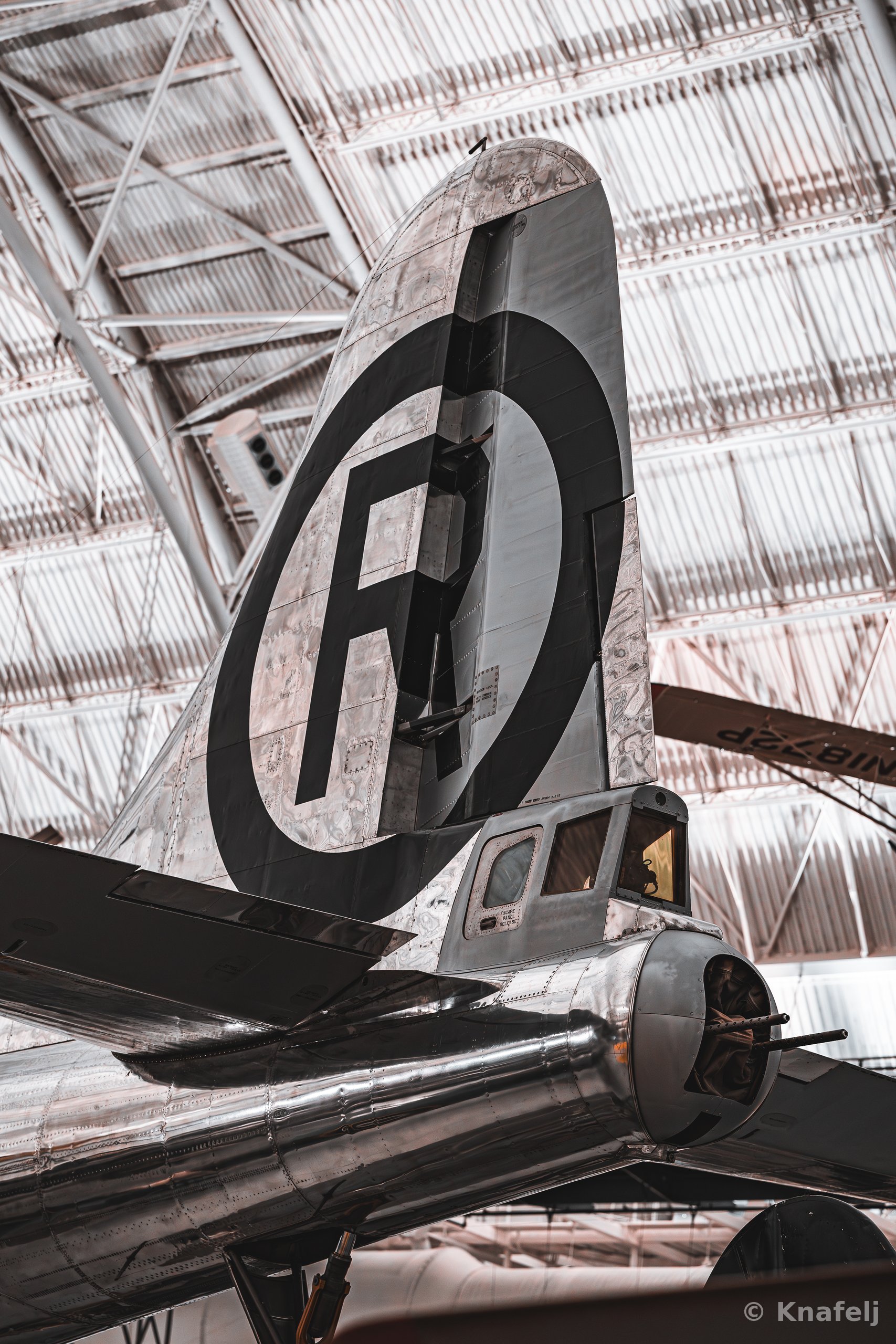 Low-angle view of a vintage silver aircraft's tail fin with a black circle and letter insignia, displayed indoors beneath a metal hangar roof.