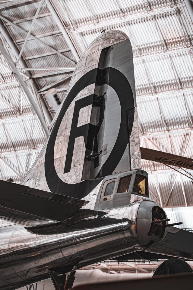 Low-angle view of a vintage silver aircraft's tail fin with a black circle and letter insignia, displayed indoors beneath a metal hangar roof.