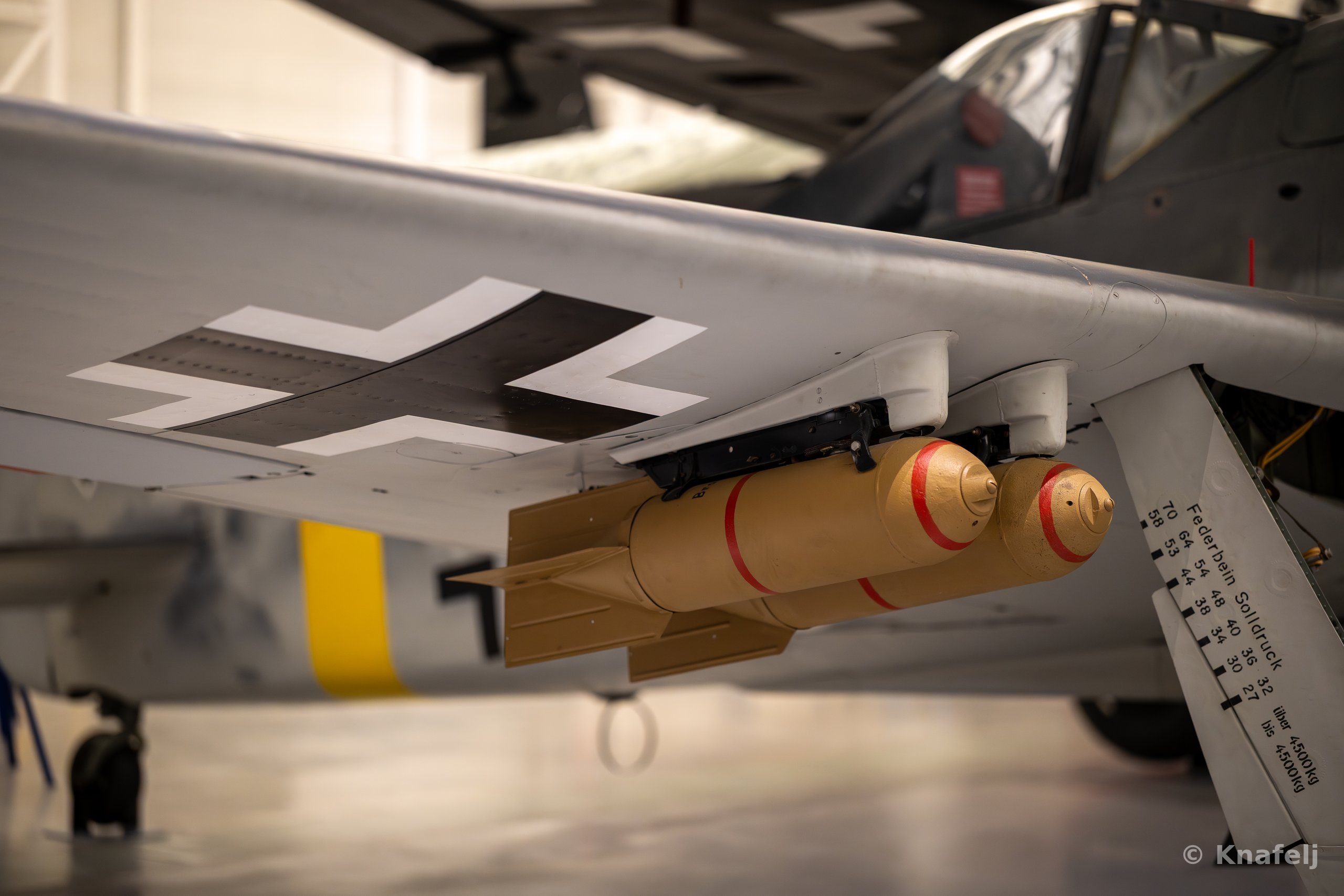 Underside of a Focke-Wulf 190 aircraft wing bearing a black and white German cross, with two tan bombs marked by red bands mounted on a pylon beneath it, displayed indoors in a museum.